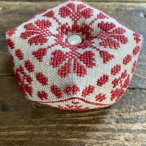 May include: A handmade, five-sided pincushion with a beige linen base and a red floral cross-stitch design. The pincushion is topped with a white button. The item is likely used for sewing and craft projects.