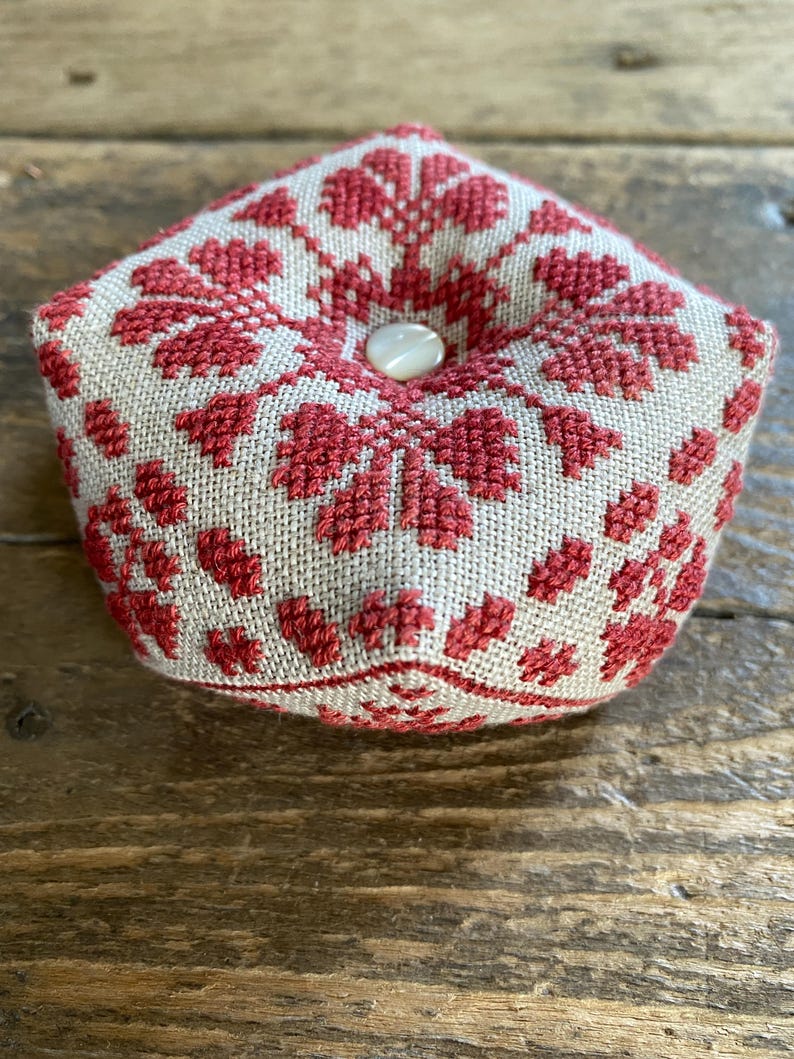 May include: A handmade, five-sided pincushion with a beige linen base and a red floral cross-stitch design. The pincushion is topped with a white button. The item is likely used for sewing and craft projects.