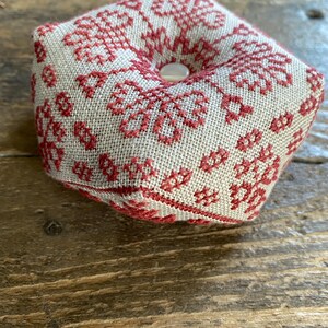 May include: A square pincushion with a beige linen fabric base, adorned with a red floral cross-stitch pattern. The center features a button. The pincushion is designed for sewing and craft projects.