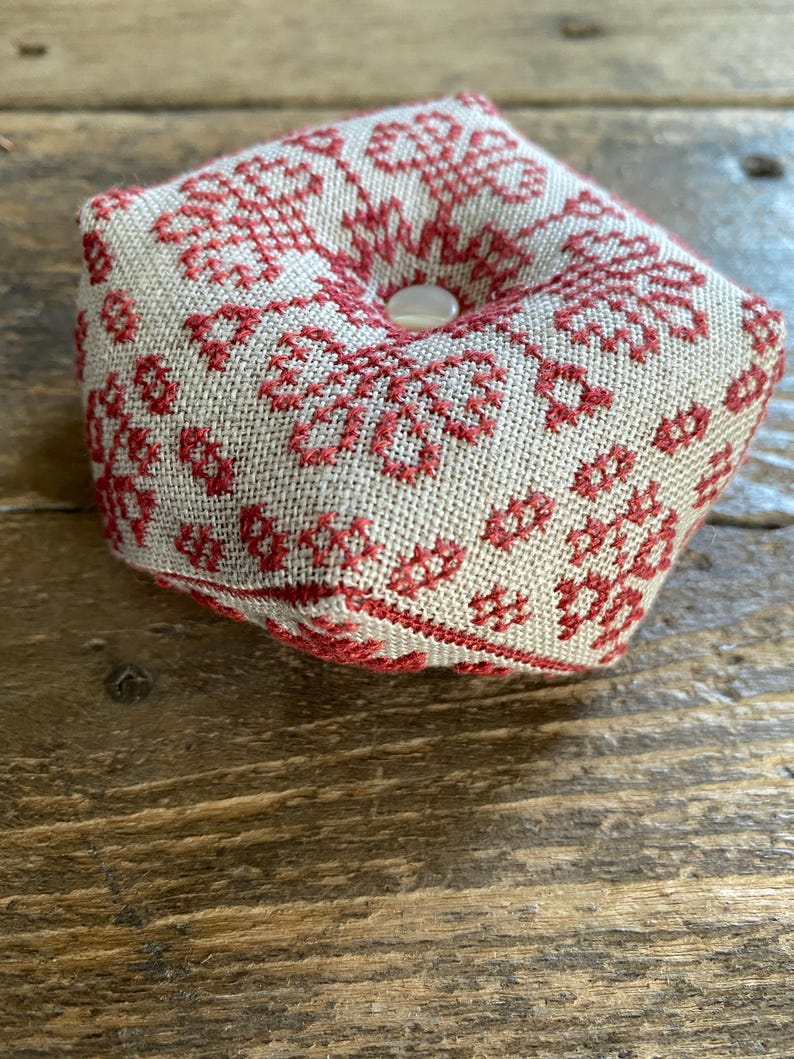 May include: A square pincushion with a beige linen fabric base, adorned with a red floral cross-stitch pattern. The center features a button. The pincushion is designed for sewing and craft projects.