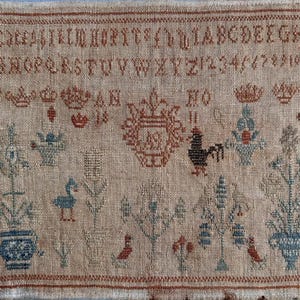 Puede incluir: Antiguo muestrario con letras bordadas, números y motivos decorativos. El muestrario presenta un escudo central, un gallo, diseños florales y varios pájaros. La paleta de colores incluye tonos de marrón, azul y rojo sobre un fondo beige.