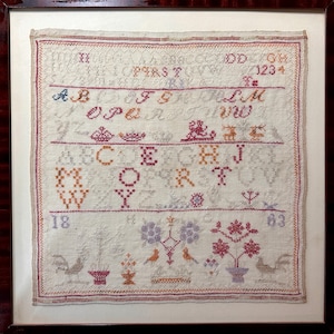 Op de afbeelding: Oude sampler in een donkerhouten lijst. De sampler toont geborduurde letters, cijfers en decoratieve motieven in verschillende kleuren, waaronder rood, oranje en blauw. Het ontwerp bevat bloemen- en vogelelementen, en de cijfers "18" en "63".