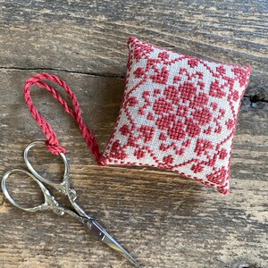 May include: A small, square, red and white embroidered sachet with a floral design and a red cord loop. Silver-colored embroidery scissors are next to the sachet on a wooden surface.