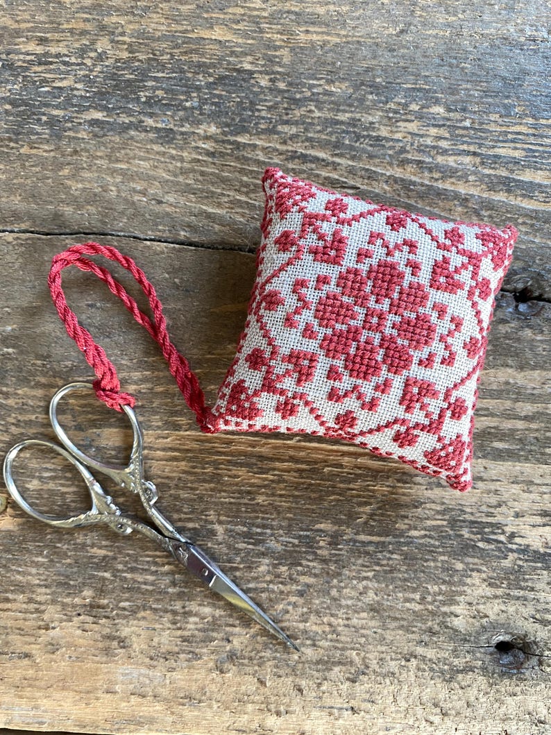 May include: A small, square, red and white embroidered sachet with a floral design and a red cord loop. Silver-colored embroidery scissors are next to the sachet on a wooden surface.