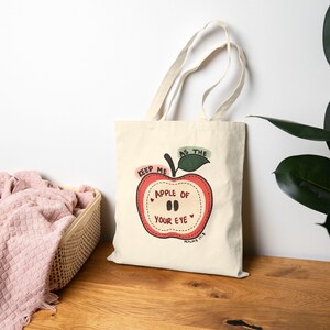 Peut inclure: Un sac fourre-tout en toile couleur crème orné d'un motif de pomme rouge à carreaux. Le texte indique "KEEP ME AS THE APPLE OF YOUR EYE" avec "PSALMS 17:8" imprimé en dessous. Le design comprend une feuille verte et des petits cœurs rouges.