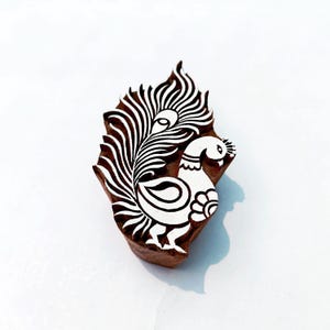 Puede incluir: Un bloque de madera con un diseño de pavo real tallado. El pavo real está representado en blanco sobre un fondo marrón, con patrones de plumas detallados. El bloque se utiliza probablemente para imprimir o estampar.