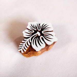 以下が含まれることがあります： 白いハイビスカスの花の模様が入った木製のブロック。花には詳細な花びらと中心の雄しべがあります。ブロックは明るい茶色の木材でできており、印刷またはスタンプに使用される可能性があります。