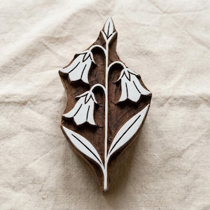 Peut inclure: Un tampon d'impression en bois brun présentant un motif floral sculpté de trois fleurs en forme de cloche sur une tige centrale avec des feuilles. Les zones sculptées sont remplies de pigment blanc, soulignant le motif botanique sur le bois.