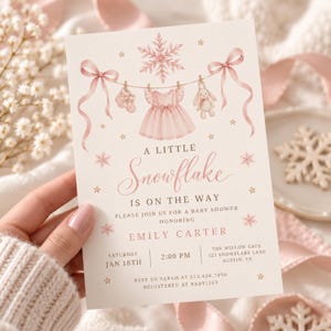 Könnte beinhalten: Eine rechteckige Babyparty-Einladungskarte mit einer rosa Aquarellillustration einer Wäscheleine mit Babyschuhen, einem Kleid und einem Plüschhasen. Der Text lautet A LITTLE Snowflake IS ON THE WAY, EMILY CARTER, JAN 18TH, 2:00 PM.