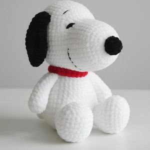 Op de afbeelding: Een handgemaakt wit gehaakt Snoopy pluche speelgoed met zwarte oren en neus, en een rode kraag. Het pluche zit met uitgestrekte benen. Het speelgoed is een zacht, knuffelbaar item.