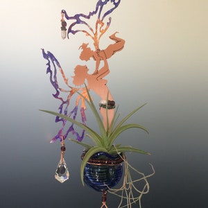 May include: A copper fairy sculpture holds a small blue glass planter with a green plant. The fairy has purple wings and is adorned with crystal accents. The sculpture hangs from a wire, creating a whimsical, decorative piece.