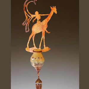 Pode incluir: Um sino de vento artesanal e caprichoso com uma silhueta de metal de uma pessoa montada numa girafa. A girafa e o cavaleiro são em tons de laranja e dourado. O sino pende de uma base de vidro e metal com elementos decorativos.