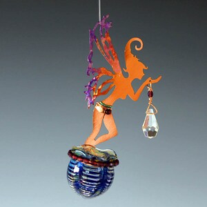 Könnte beinhalten: Kupferfarbenes Feenornament mit lila Flügeln und einer Kristalllaterne. Die Fee sitzt auf einer blau-goldenen Glaskugel. Das Ornament wird von einem dünnen Draht gehalten und schafft ein skurriles Dekorationsobjekt.