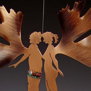 Pode incluir: Um recorte de metal de cobre de duas figuras com asas, de frente uma para a outra. As figuras estão de mãos dadas e têm um bracelete de contas em um pulso.
