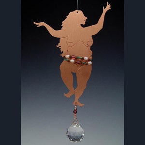 May include: Copper metal silhouette of a dancing figure with arms raised. The figure has a beaded belt with white and pink beads. A clear crystal ball hangs below the figure. The silhouette is suspended by a thin wire.