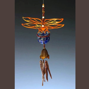 May include: A handcrafted wind chime with a dragonfly design. The dragonfly has orange wings and a blue and clear glass body. The chime includes a bell-shaped bottom and dangling wooden pieces. The wind chime is suspended by a thin string.
