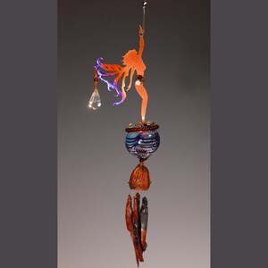 Pode incluir: Um sino de vento artesanal com uma silhueta cor de cobre de uma mulher com os braços estendidos. O sino de vento inclui um globo de vidro azul e branco, um pingente de cristal e vários elementos de madeira e pedra. O design geral tem cerca de 46 cm de comprimento.