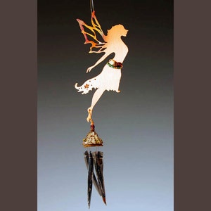 May include: A copper fairy wind chime with a delicate silhouette of a fairy with wings and a star on her dress. The fairy is suspended from a small, ornate, gold-colored bell with a red bead at the top. The chime has four long, thin, silver-colored metal pieces that hang from the bell.