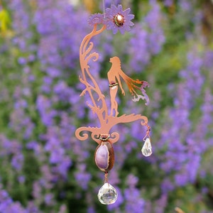 May include: Copper metal wind chime featuring a silhouette of a woman standing on a tree branch with a purple flower at the top. The chime has a purple and brown seed pod and a crystal prism hanging from it.