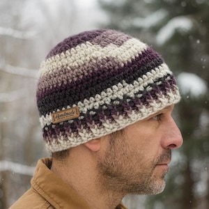 Może przedstawiać: Ręcznie robiona czapka z szydełka w odcieniach fioletu, szarości i kremu. Czapka ma poziome paski i małą brązową skórzaną metkę z napisem "Handmade". Czapka została zaprojektowana z myślą o cieple i stylu.