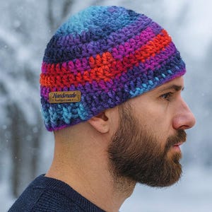 May include: A handmade, colourful crochet beanie in shades of blue, purple, and red. The beanie has a striped pattern and a small brown tag that reads "Handmade". The hat is designed for cold weather.