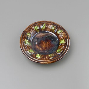 May include: A round ceramic dish with a glossy dark brown glaze. The rim features a raised relief pattern of small, textured green leaves arranged in a circular sequence around the center, which has a smooth, deep brown finish.