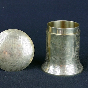 Może przedstawiać: Srebrny, cylindryczny pojemnik z pokrywką. Pokrywka jest okrągła z wytrawionym kwiatowym wzorem. Pojemnik ma rozszerzoną podstawę i wytrawione wzory kwiatowe. Pojemnik jest otwarty, odsłaniając puste wnętrze.