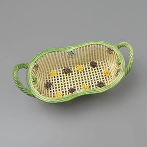 Pode incluir: Uma cesta oval de cerâmica com corpo em treliça cor de creme e borda trançada verde. Duas alças trançadas verdes correspondentes estendem-se dos lados. Pequenos detalhes florais coloridos em amarelo, marrom e verde estão na treliça.