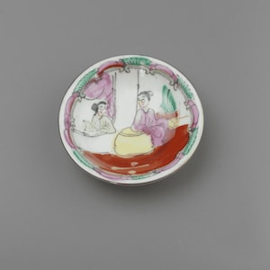 Peut inclure: Petite assiette ronde en céramique peinte à la main, ornée d'une scène détaillée de personnages dans une pièce. Le bord est décoré de motifs floraux verts et roses. Les personnages sont vêtus de vêtements traditionnels.