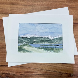 May include: Watercolour landscape art print depicting a mountain range with snow-covered peaks, a lake, and green hills beneath a blue sky. The artwork is framed in a white mount and presented with a white envelope.