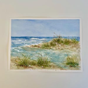 May include: Watercolour painting of a coastal scene. The artwork depicts a sandy shore with tufts of green grass, clear blue water, and a light blue sky. The waves are crashing in the distance, creating a serene and natural landscape.