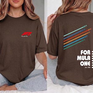 Pu&ograve; includere: Una t-shirt a maniche corte marrone scuro con un piccolo logo rosso Formula One sul petto. Il retro mostra una grafica con cinque linee diagonali colorate con icone di auto da corsa, il testo FORMULA ONE e la scritta est. 1950.
