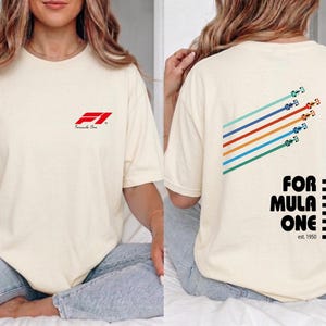 Pu&ograve; includere: Una t-shirt a maniche corte color crema con un piccolo logo rosso Formula 1 sul petto. Il retro mostra una grafica di cinque auto da corsa colorate con linee di velocit&agrave; diagonali e il testo Formula One est. 1950 con un bordo a scacchi.