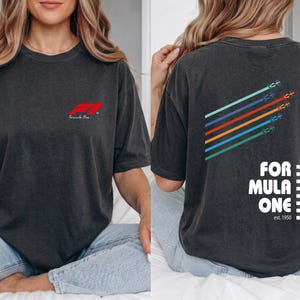 Pu&ograve; includere: Una t-shirt grigio antracite con un piccolo logo rosso Formula One sul petto. Il retro presenta una grafica con cinque linee diagonali colorate con icone di auto da corsa, il testo FORMULA ONE, un motivo a scacchi e la scritta est. 1950.