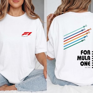 Pu&ograve; includere: Una t-shirt bianca con un piccolo logo rosso Formula One sul petto a sinistra. Il retro mostra una grafica di cinque auto da corsa colorate con linee di velocit&agrave; diagonali, il testo "FORMULA ONE" in nero e un motivo a scacchi verticale.