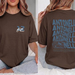 May include: Espresso-coloured t-shirt with blue text and graphics. The front features the number "32" and the name "Kimi" in a script font. The back displays the name "Antinelli" in a wavy design, along with a car graphic.