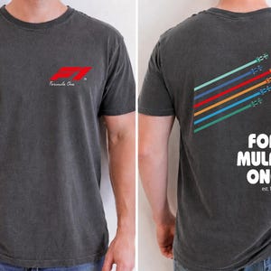 Pu&ograve; includere: Una t-shirt grigio antracite a maniche corte con un piccolo logo rosso Formula One sul petto. Il retro mostra una grafica di sei auto da corsa colorate con linee di velocit&agrave; diagonali e il testo Formula One est. 1950 con un bordo a scacchi.