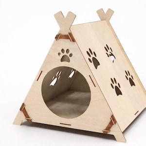 Puede incluir: Un tipi de madera clara para mascotas con recortes de huellas de patas y una entrada circular. El tipi tiene forma triangular con base plana y se sujeta con correas de cuero.