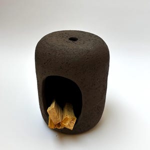 Peut inclure: Un porte-encens en céramique brun foncé et texturé, avec un dessus arrondi et une grande ouverture. À l'intérieur de l'ouverture se trouvent plusieurs morceaux de bois brun clair. Le porte-encens a un petit trou sur le dessus.