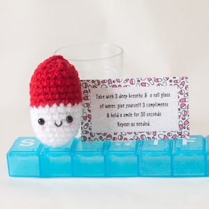 May include: A crocheted pill-shaped plush toy with a red cap and a white body. The toy has a happy face and is sitting in a blue pill organizer. A white card with pink and red pill designs and text that reads "Take with 3 deep breaths & a tall glass of water. give yourself 3 compliments & hold a smile for 30 seconds. Repeat as needed." is behind the toy.