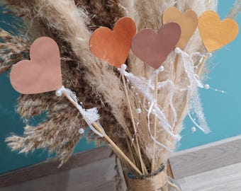 Puede incluir: Un ramo de hierba de la Pampa seca y decoraciones en forma de corazón. Los corazones son en tonos naranja y marrón, unidos a palitos de madera con encaje y perlas. El ramo está en un jarrón de cristal.