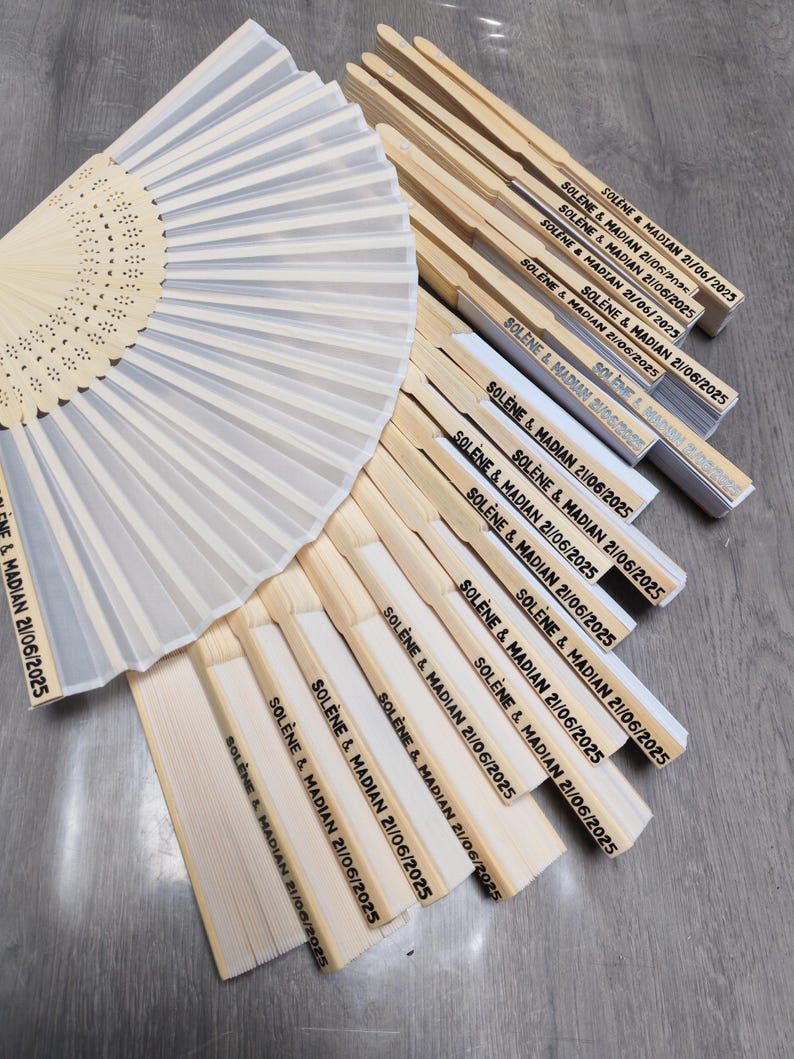 Op de afbeelding: Een verzameling gepersonaliseerde vouwventilatoren. Sommige ventilatoren zijn open en tonen een geplooide, lichtgekleurde stof. De gesloten ventilatoren zijn gemaakt van licht hout en hebben de tekst "SOL&Egrave;NE & MADIAN 21/06/2025".