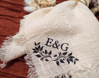 Op de afbeelding: Crèmekleurig linnen servet met gerafelde randen, geborduurd met de initialen "E&G" en een zwart bloemmotief. Een gouden ring houdt het servet vast, met gedroogde bloemen op de achtergrond. Het servet ligt op een roestkleurig oppervlak.