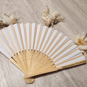 Op de afbeelding: Een wit en goudkleurige vouwventilator met een houten handvat, met de inscriptie "Elodie & Gautier 31.05.2025". De ventilator heeft wit papier en gouden accenten. Gedroogde bloemstukken zijn rond de ventilator geplaatst.