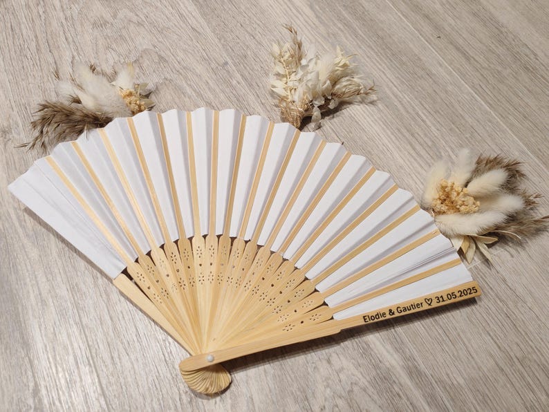 Op de afbeelding: Een wit en goudkleurige vouwventilator met een houten handvat, met de inscriptie "Elodie & Gautier 31.05.2025". De ventilator heeft wit papier en gouden accenten. Gedroogde bloemstukken zijn rond de ventilator geplaatst.