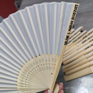 Op de afbeelding: Een witte en lichtgrijze vouwventilator met een bamboe handvat. De ventilator heeft een delicaat, geplooid ontwerp met verticale strepen. De tekst "SOL&Egrave;NE & MADIAN 21/06/2025" is op de zijkant gedrukt.
