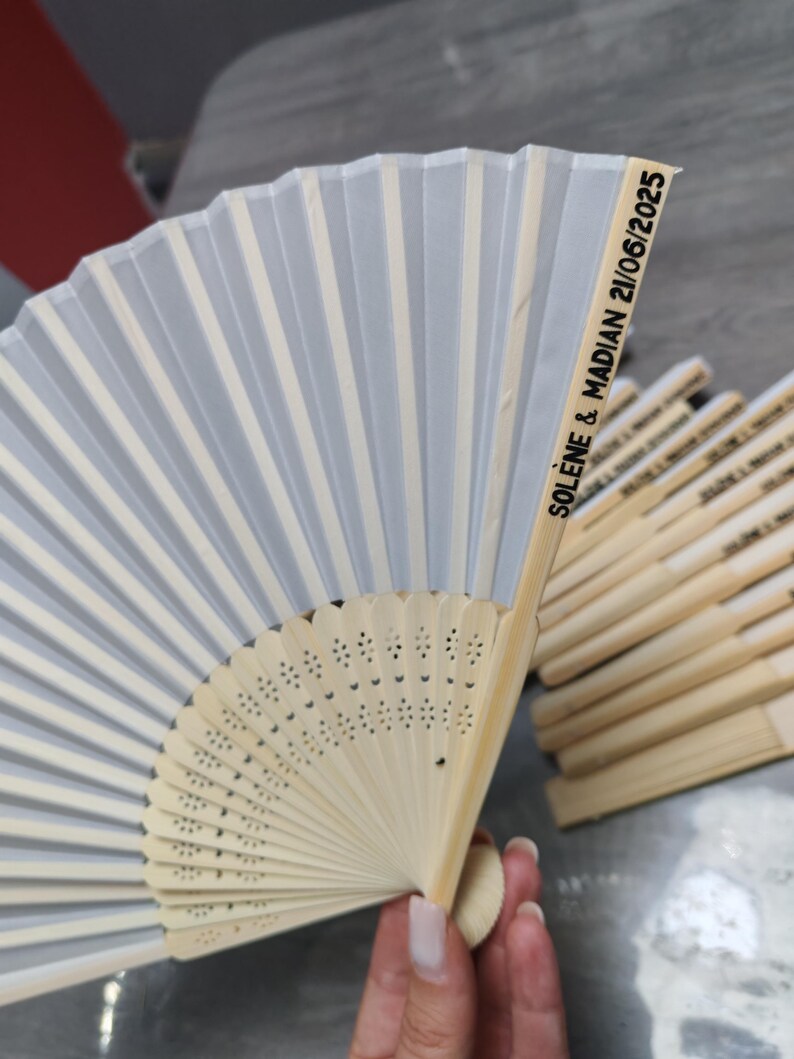 Op de afbeelding: Een witte en lichtgrijze vouwventilator met een bamboe handvat. De ventilator heeft een delicaat, geplooid ontwerp met verticale strepen. De tekst "SOL&Egrave;NE & MADIAN 21/06/2025" is op de zijkant gedrukt.