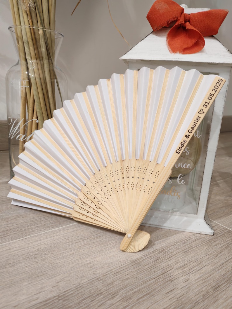 Op de afbeelding: Een witte en natuurlijk houten vouwventilator met de tekst "Elodie & Gautier 31.05.2025". De ventilator is open en toont witte papieren panelen en natuurlijke houten ribben. De ventilator ligt op een licht houten oppervlak.