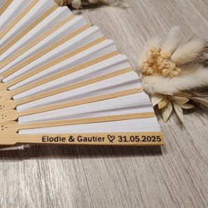 Op de afbeelding: Een witte en bamboe handventilator met de namen "Elodie & Gautier" en de datum "31.05.2025" gegraveerd op het handvat. De ventilator is gedeeltelijk open en onthult wit papier en bamboeribben. Een klein bloemstuk ligt naast de ventilator.