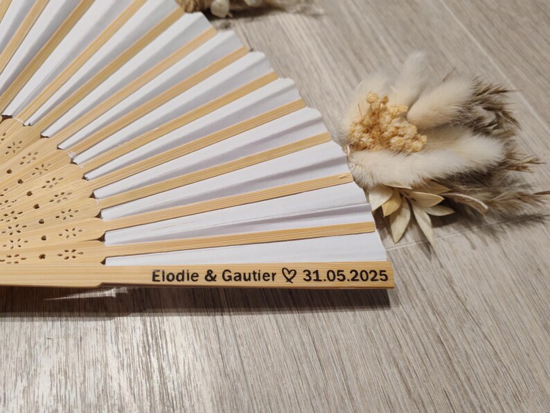 Op de afbeelding: Een witte en bamboe handventilator met de namen "Elodie & Gautier" en de datum "31.05.2025" gegraveerd op het handvat. De ventilator is gedeeltelijk open en onthult wit papier en bamboeribben. Een klein bloemstuk ligt naast de ventilator.
